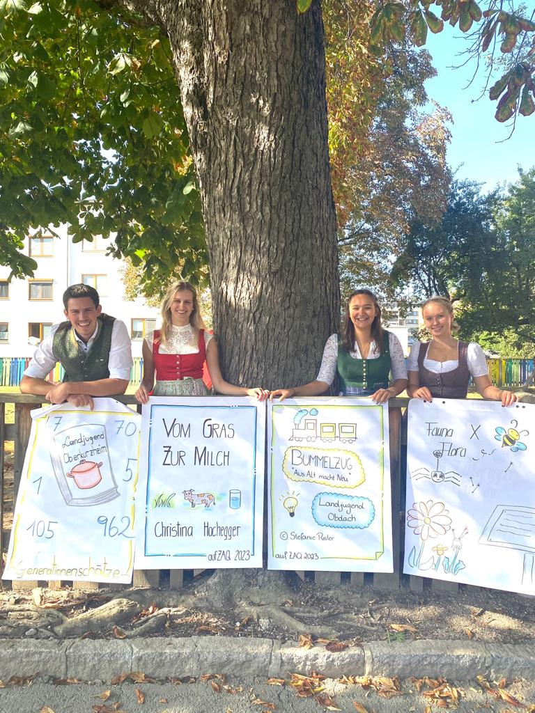 Vier junge Menschen in Tracht stehen unter einem Baum. Jede/r hält ein Poster mit ihrer Projektidee in der Hand